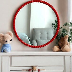 May include: A round mirror with a red beaded frame hangs above a white dresser. A plush teddy bear wearing a blue blanket sits on the left, and another teddy bear is on the right. A plant is next to the right teddy bear.