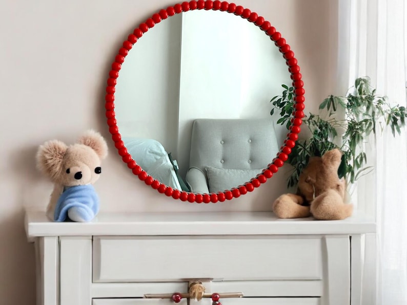 May include: A round mirror with a red beaded frame hangs above a white dresser. A plush teddy bear wearing a blue blanket sits on the left, and another teddy bear is on the right. A plant is next to the right teddy bear.