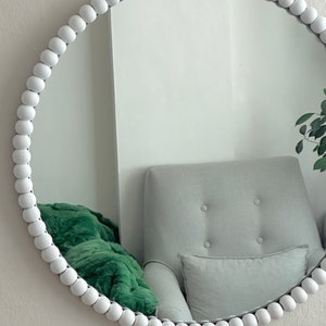 May include: A round white beaded mirror. The mirror's frame is made of small, evenly spaced white beads. The reflection shows a gray armchair, a green blanket, and a white wall. The mirror is a decorative home decor item.