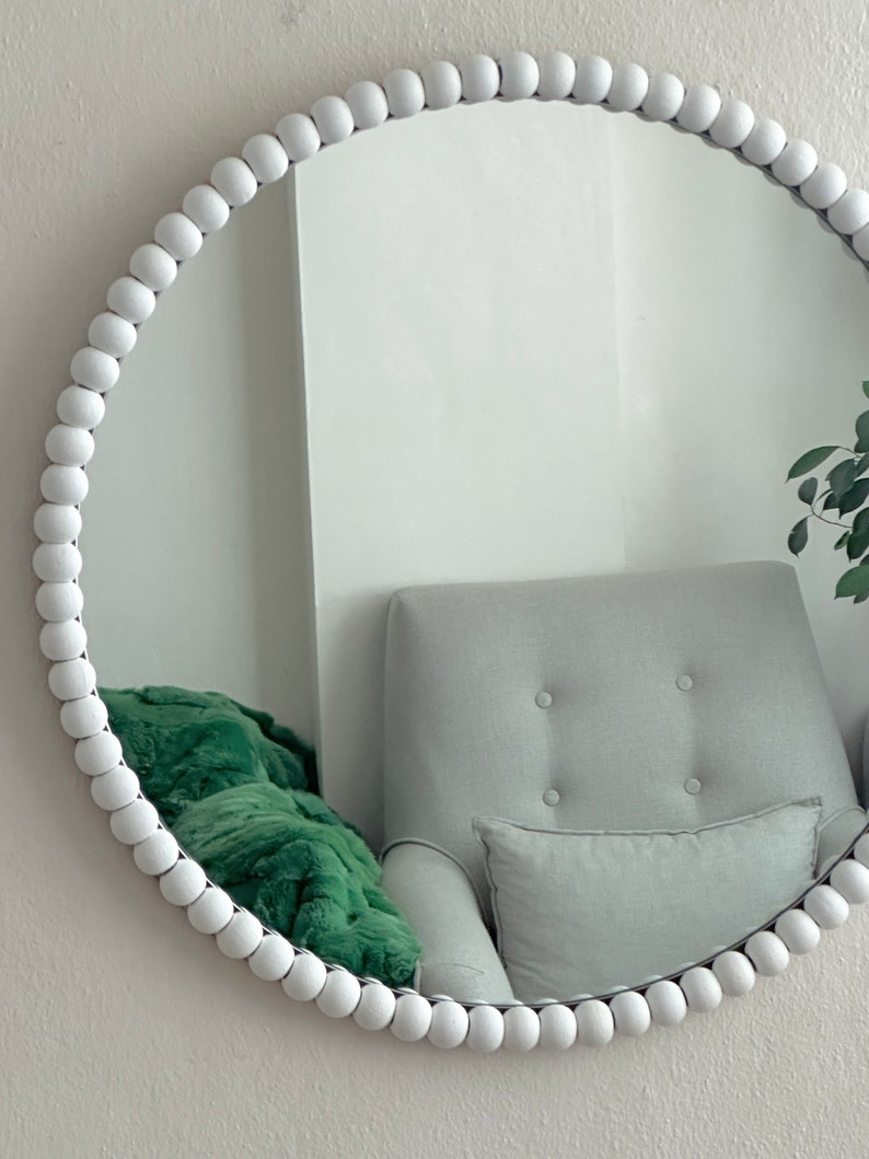 May include: A round white beaded mirror. The mirror's frame is made of small, evenly spaced white beads. The reflection shows a gray armchair, a green blanket, and a white wall. The mirror is a decorative home decor item.