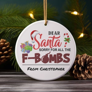 May include: A white ceramic Christmas ornament with the text "DEAR Santa, SORRY FOR ALL THE F-BOMBS FROM Christopher." The ornament features a Santa hat, wrapped gifts, and a candy cane.