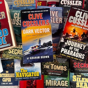 May include: A collection of paperback books by Clive Cussler and other authors. The books feature colorful covers with titles like "Dark Vector," "Journey of the Pharaohs," and "The Navigator." The books are from the Numa Files and Fargo Adventure series.