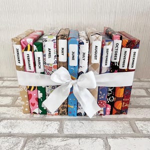 May include: A collection of twelve books, each wrapped in patterned paper and labeled with a month from January to December. A white ribbon with a bow secures the books together. The books are arranged upright.