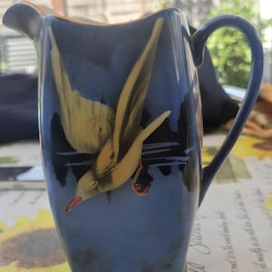 May include: A blue ceramic jug featuring a hand-painted yellow bird in flight over dark water. The jug has a curved handle, a pouring spout, and a gold-toned rim. The design includes black accents and a small red detail on the bird's foot.