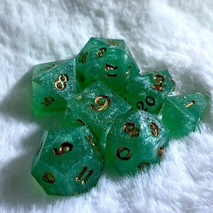 May include: A set of seven polyhedral dice in various shapes, perfect for tabletop games. The dice are a translucent green with gold-colored numbers. The dice are resting on a white, fluffy surface.