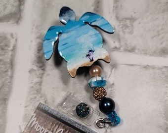 May include: A blue and white painted wooden turtle with a beach scene on its shell. The turtle is attached to a beaded lanyard with a clear badge holder. The lanyard has blue, white, gold, and black beads. The badge holder has a small blue and black sparkly embellishment. The turtle is a charm for a badge holder or key chain. The card below the turtle reads "Moore Naturals & Crafts Handmade Natural Face & Body Products, Trendy Crafts & Home Decor tena & stephanie moore moorenaturalsandcrafts@gmail.com"