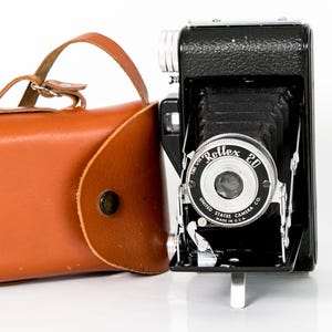 May include: Vintage black Rollex 20 camera with a brown leather case. The camera has a silver lens and the words "Rollex 20" are printed on the front. The case has a handle and a strap.