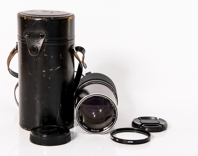 K&ouml;nnte beinhalten: Ein schwarzes Kameraobjektiv mit Objektivdeckel, Filter und einer schwarzen Ledertasche mit Riemen. Das Objektiv ist silber und schwarz mit dem Text "Olympus". Die Tasche ist zylindrisch.
