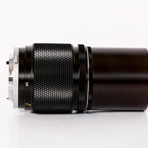 K&ouml;nnte beinhalten: Ein schwarzes und silbernes Kameraobjektiv mit strukturiertem Griff und dunkelbraunem Geh&auml;use. Das Objektiv hat Markierungen und Zahlen, darunter "2.5" und "Lens Made in Japan". Vor wei&szlig;em Hintergrund.