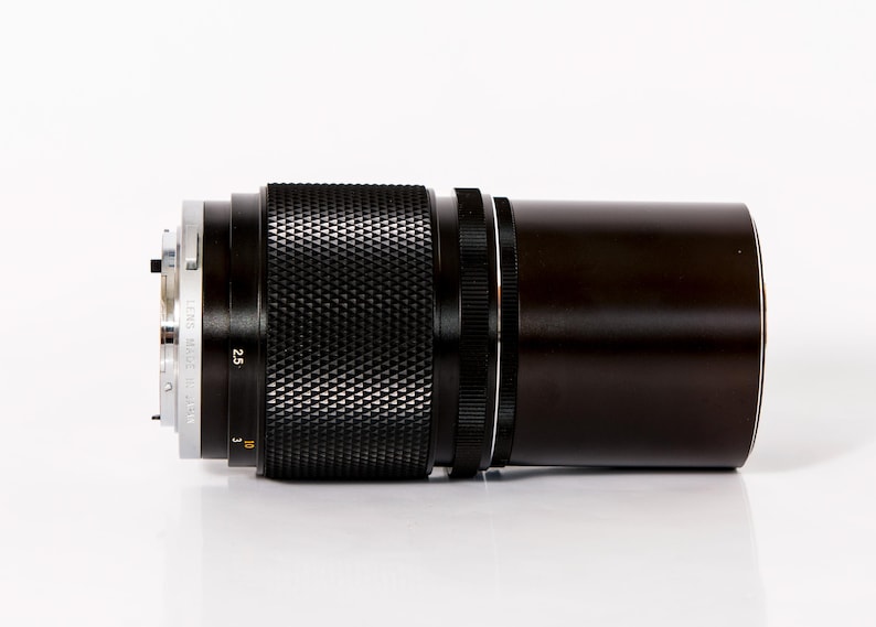 K&ouml;nnte beinhalten: Ein schwarzes und silbernes Kameraobjektiv mit strukturiertem Griff und dunkelbraunem Geh&auml;use. Das Objektiv hat Markierungen und Zahlen, darunter "2.5" und "Lens Made in Japan". Vor wei&szlig;em Hintergrund.