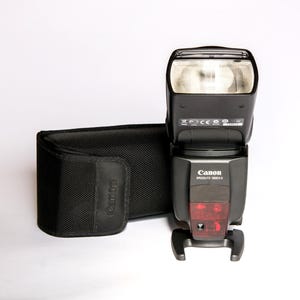 May include: A black Canon Speedlite 580EX II flash unit and a matching black carrying case. The flash has a rectangular head with a clear front and a red panel. The Canon logo is visible on the front of the flash.