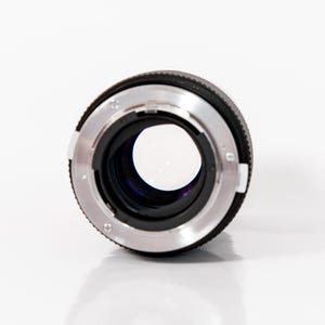 K&ouml;nnte beinhalten: Nahaufnahme eines Kameraobjektivs. Es hat ein schwarzes Geh&auml;use mit einem silberfarbenen Metallring. Das Objektiv befindet sich vor einem wei&szlig;en Hintergrund, wobei das Glas das Licht reflektiert.