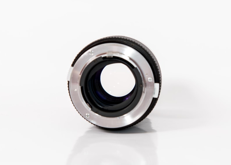 K&ouml;nnte beinhalten: Nahaufnahme eines Kameraobjektivs. Es hat ein schwarzes Geh&auml;use mit einem silberfarbenen Metallring. Das Objektiv befindet sich vor einem wei&szlig;en Hintergrund, wobei das Glas das Licht reflektiert.