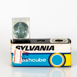 May include: A Sylvania Flashcube sits atop its original box. The flashcube is a clear, square-shaped device with a silver interior. The box is white with blue and yellow accents and the Sylvania logo.