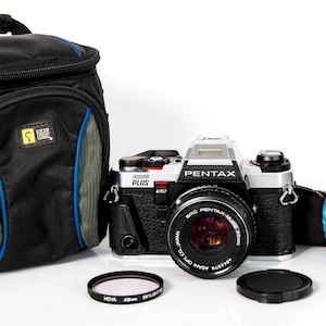 May include: A black and silver Pentax camera with a lens, lens cap, and filter. A black camera bag with blue accents is on the left. The camera strap is black with blue accents.