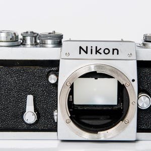 May include: A vintage Nikon film camera body featuring a silver top and bottom plate with a black textured grip. The front displays the Nikon logo, a circular lens mount, a self-timer lever, and various metallic dials and buttons on the top.