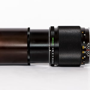 K&ouml;nnte beinhalten: Ein schwarzes und braunes Kameraobjektiv mit einem silbernen Ring und einem strukturierten Griff. Das Objektiv hat eine 200-mm-Markierung und verschiedene numerische Indikatoren. Das Objektiv liegt horizontal auf einer wei&szlig;en Oberfl&auml;che.