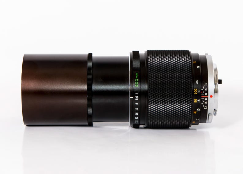 K&ouml;nnte beinhalten: Ein schwarzes und braunes Kameraobjektiv mit einem silbernen Ring und einem strukturierten Griff. Das Objektiv hat eine 200-mm-Markierung und verschiedene numerische Indikatoren. Das Objektiv liegt horizontal auf einer wei&szlig;en Oberfl&auml;che.