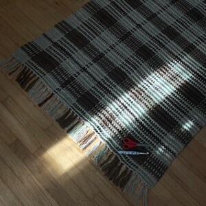 May include: A handmade, rectangular crochet blanket with a plaid pattern in shades of brown, grey, and light blue. The blanket has a fringed edge and is lying on a wooden floor. A pair of red scissors and a crochet hook are on the blanket.
