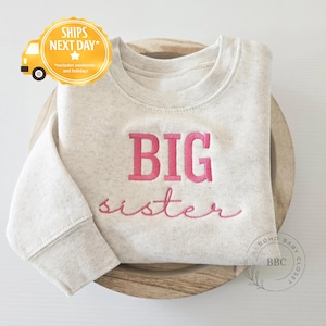 May include: A light gray sweatshirt with the words "BIG sister" embroidered in pink. The sweatshirt is displayed on a wooden tray. The image also includes a yellow circle with a truck graphic and the text "SHIPS NEXT DAY".