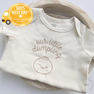 May include: Cream-colored baby onesie with the embroidered text "our little dumpling" and a dumpling graphic. The onesie is made by Gerber and is displayed in a wooden bowl. The onesie is a baby clothing item.