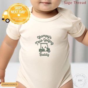 May include: Cream-colored baby onesie with the embroidered text "Grumpy's New Golfing Buddy" and a golf cart graphic in green. The onesie has short sleeves and a round neckline. The text "Ships Next Day" is also visible.