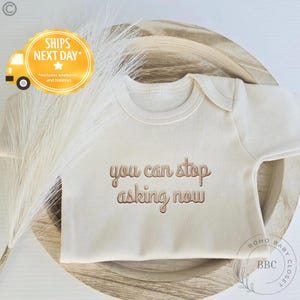 May include: Cream-colored baby onesie with the phrase "you can stop asking now" in a brown, embossed font. The onesie is displayed on a wooden surface with dried ornamental grass. The Boho Baby Closet logo is visible.