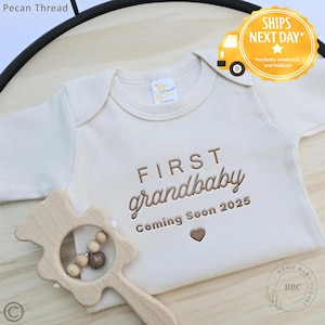 May include: Cream-colored baby onesie with the embroidered text "FIRST grandbaby Coming Soon 2025" and a heart symbol. A wooden rattle is in the foreground. The image also includes the text "SHIPS NEXT DAY".