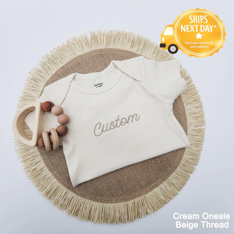 A cream-colored onesie with the word "Custom" embroidered on it, placed on a round beige fringed placemat.
