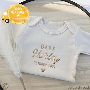 May include: Cream-colored baby onesie with the name "Harley" embroidered in a light brown thread, along with "BABY" and "OCTOBER 2024". A small heart is embroidered below the date. The onesie is a long-sleeved style.