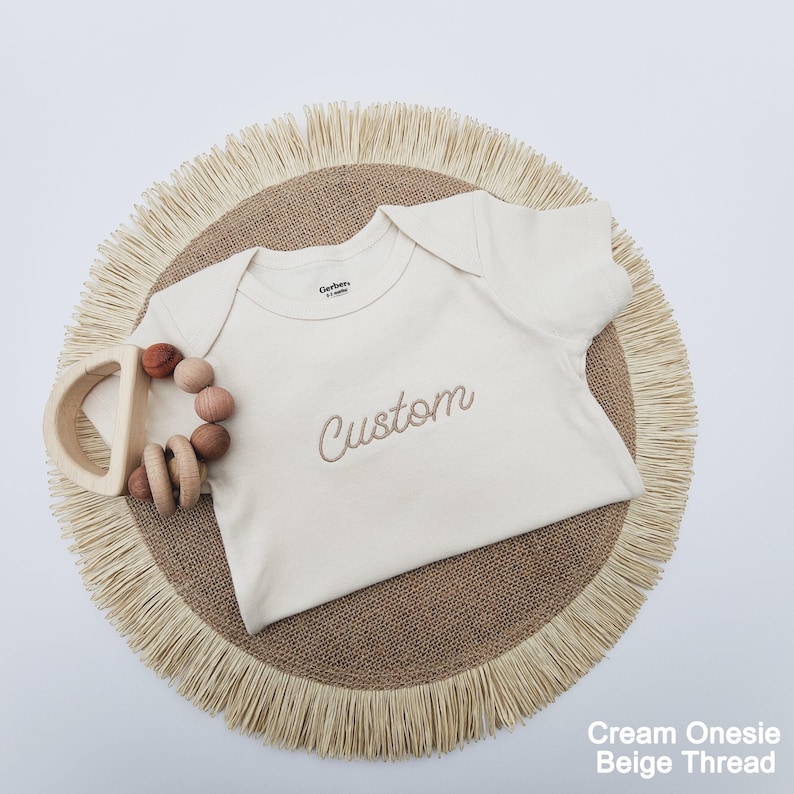 A cream-colored onesie with the word "Custom" embroidered on it, placed on a round straw mat with fringed edges.