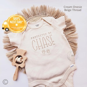 May include: Cream-colored baby onesie with "PROTECTED BY CHASE" embroidered in beige thread, along with paw prints. A wooden rattle is next to the onesie. The text "Cream Onesie Beige Thread" is visible.