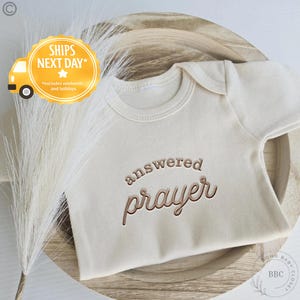May include: A cream-colored baby onesie with the words "answered prayer" embroidered in a brown script. The onesie is displayed on a wooden tray with decorative elements. The image also includes a logo and text.