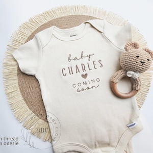 A baby onesie with the text "baby CHARLES" and a crocheted teddy bear are displayed on a round straw mat.