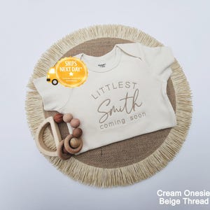 A cream-colored onesie with the text "LITTLE Smith coming soon" printed on it, along with a wooden teething ring and a fringed placemat.