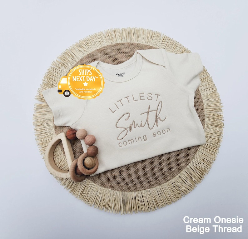 A cream-colored onesie with the text "LITTLE Smith coming soon" printed on it, along with a wooden teething ring and a fringed placemat.