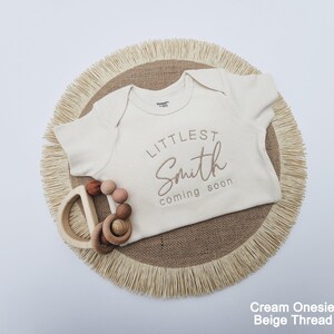 A cream-colored onesie with the text "LITTLE Smith coming soon" printed on it, along with a wooden teething ring and a beige fringed placemat.