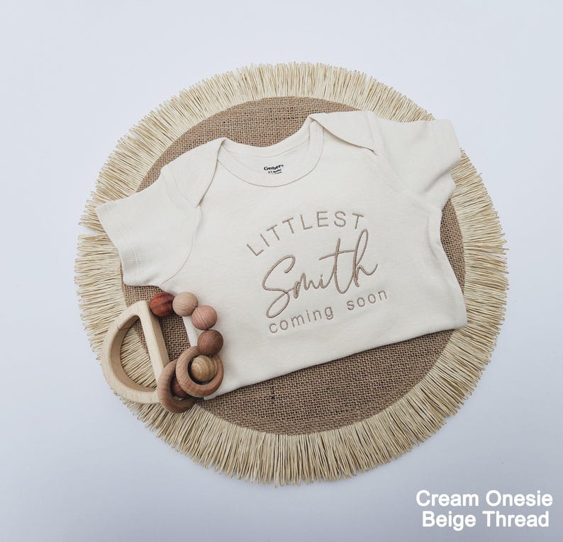 A cream-colored onesie with the text "LITTLE Smith coming soon" printed on it, along with a wooden teething ring and a beige fringed placemat.