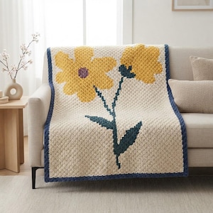 May include: A cream-coloured crochet blanket featuring a large yellow flower with a purple centre and a second yellow flower bud on a dark teal stem with leaves. The blanket has a dark teal border and a textured, square-stitch pattern throughout.