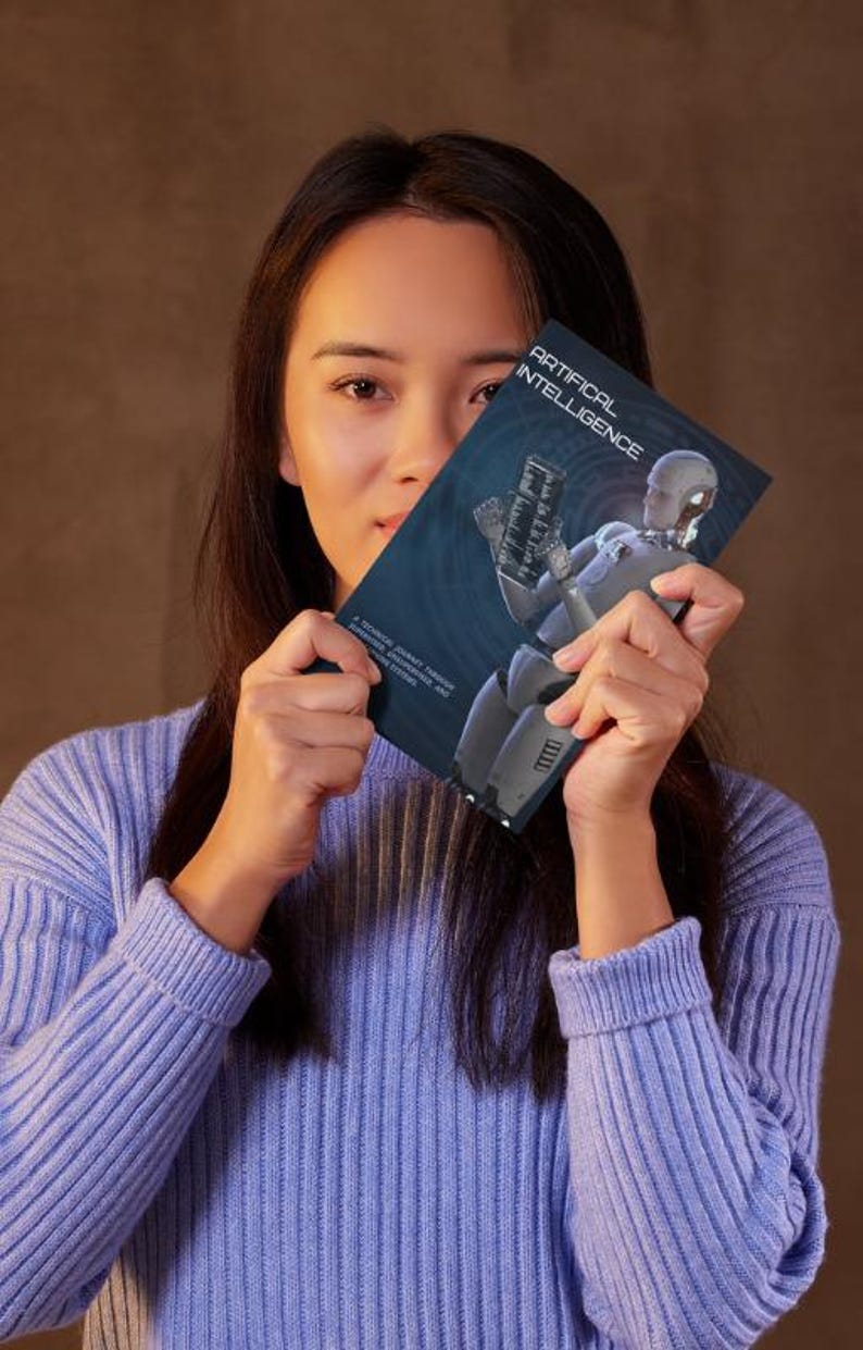 K&ouml;nnte beinhalten: Eine Person h&auml;lt ein Buch mit dem Titel "ARTIFICIAL INTELLIGENCE". Das Buchcover zeigt eine futuristische Roboterillustration in Grau- und Blaut&ouml;nen. Die Person tr&auml;gt einen hellblauen Pullover.