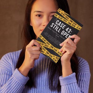 May include: A person holding a book titled "CASE 43: STILL OPEN" with yellow and black crime scene tape design. The book's cover is black with white text. The person is wearing a light blue ribbed sweater.