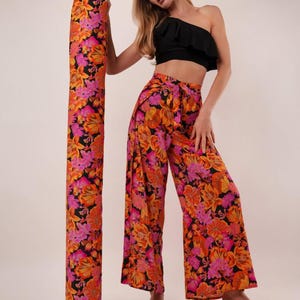 May include: A woman wearing a black one-shoulder crop top and wide-leg pants with a vibrant floral print in shades of orange, pink, and black. A matching fabric roll stands beside her. The outfit is paired with black sandals.