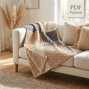Op de afbeelding: Een gebreide plaid met een kleurblokontwerp in tinten beige, crème en leigrijs met gevarieerde gestructureerde steekpatronen. De deken ligt over een bank en bevat een digitaal label met de tekst PDF Pattern.