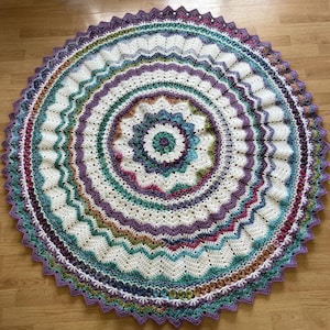 May include: A circular crochet blanket featuring a mandala-inspired pattern with concentric rings of white, purple, teal, and green yarn. The design incorporates intricate stitch textures, wavy borders, and a scalloped edge in deep purple.