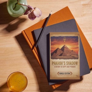 Puede incluir: Un libro titulado "Pharoh's Shadow" con una ilustración de pirámides, sobre una pila de cuadernos naranjas y grises. Un lápiz negro cruza los cuadernos. También hay un vaso de líquido amarillo y un pequeño jarrón verde con flores moradas.