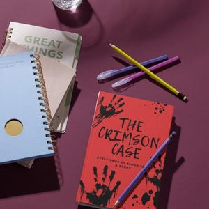 May include: A collection of stationery on a purple surface, including a red book titled "The Crimson Case" with handprints and the text "Every drop of blood tells a story." There are also notebooks, pencils, and a glass of water.