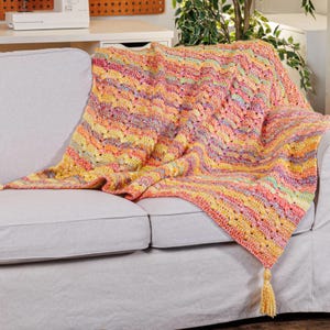 May include: A handmade crochet throw blanket featuring a vibrant variegated yarn pattern in shades of pink, orange, yellow, and light green. The blanket has a textured wavy stitch design and is finished with a single decorative tassel at the corner.
