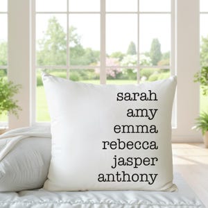 May include: White decorative pillow with the names Sarah, Amy, Emma, Rebecca, Jasper, and Anthony printed in black. The pillow is on a white surface in front of a window with plants.