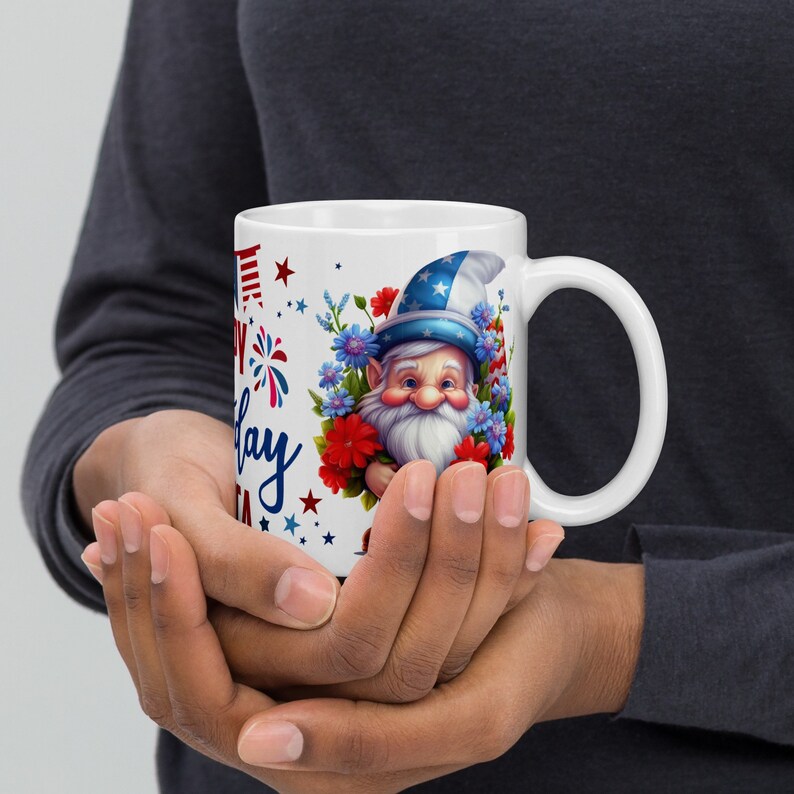 Op de afbeelding: Witte keramische koffiemok met een rood-wit-blauwe dwerg die een bos bloemen vasthoudt. De tekst "Happy 4th of July" staat op de mok gedrukt.