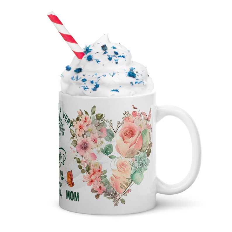 May include: White ceramic mug with a green floral heart design and the text "To a very special Mom". The mug has a red and white striped straw and whipped cream with blue sprinkles on top.